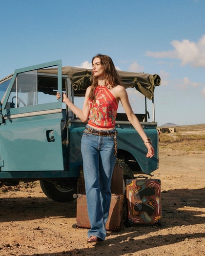 Woman wearing an ETRO printed halter top with flared jeans beside a vintage off-road vehicle in a desert landscape.
