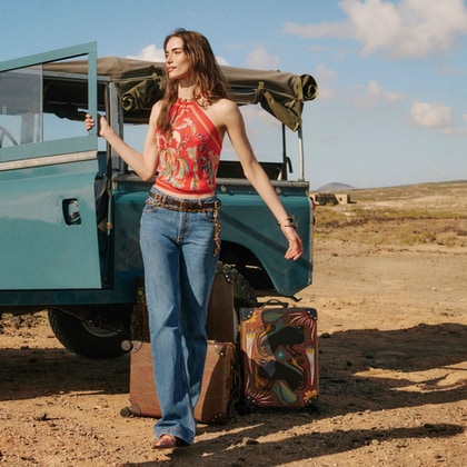 Woman wearing an ETRO printed halter top with flared jeans beside a vintage off-road vehicle in a desert landscape.