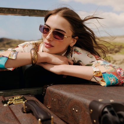 Woman wearing an Etro paisley-print shirt and matching scarf, accessorised with sunglasses and jewellery, leaning on vintage luggage outdoors.