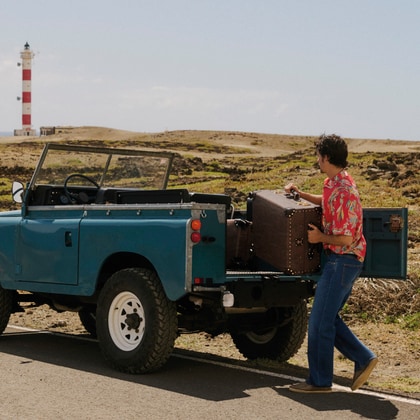 Man wearing an ETRO printed shirt with jeans beside a vintage off-road vehicle in a desert landscape.