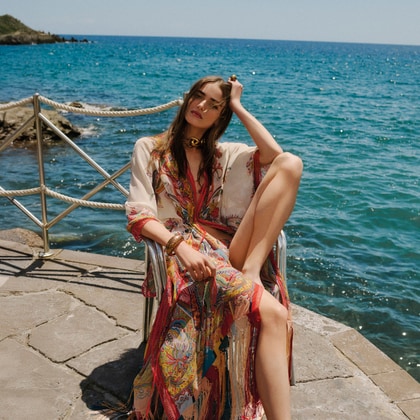 Woman wearing a flowing Etro paisley beach cover-up, seated by the seaside on a sunny day.
