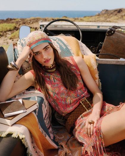 Model wearing an Etro sleeveless paisley-print dress with a matching headscarf, accessorized with statement jewelry and a woven belt, reclining in an open-top vehicle by the coast.