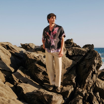 Man wearing an Etro printed short-sleeve shirt and relaxed trousers, standing on coastal rocks.