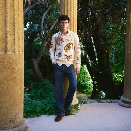 Model wearing an Etro patterned shirt and jeans, standing between stone columns in a lush garden.