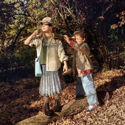 Two children wearing ETRO outfits explore a woodland setting, using a magnifying glass and a telescope among fallen leaves and trees.