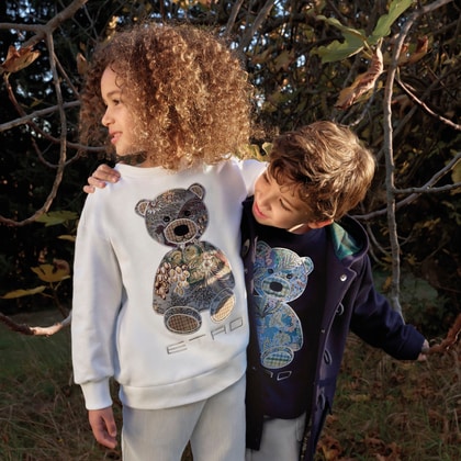 Two children wearing ETRO Kids sweatshirts featuring colorful teddy bear motifs stand together outdoors, surrounded by sunlit foliage.