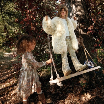 Two children wearing ETRO Kids outfits play outdoors beside a rope swing, featuring paisley prints, flowing silhouettes and textured outerwear in a woodland setting.