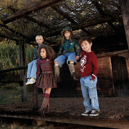 Four children wearing ETRO Kids outfits pose outdoors on a wooden structure, showcasing plaid, paisley and graphic designs in a nature-inspired setting.