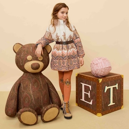 Young girl wearing an Etro Kids outfit with a patterned dress, orange tights, and boots, posing beside a large paisley teddy bear and decorative block.