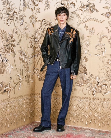 Model wearing a dark leather jacket with floral embroidery over a patterned shirt and blue trousers.