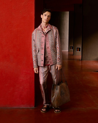 A male model in a coordinated Etro ensemble featuring a paisley-patterned jacket and trousers, layered over a matching shirt, accessorized with a choker necklace, tote bag, and sandals.