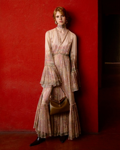 A woman in a flowing, bohemian-style Etro dress with sheer sleeves, ruffle details, and a paisley print, accessorized with statement earrings and a brown handbag.