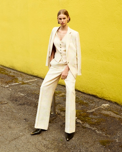 A woman poses against a yellow wall wearing a chic cream-colored three-piece suit.