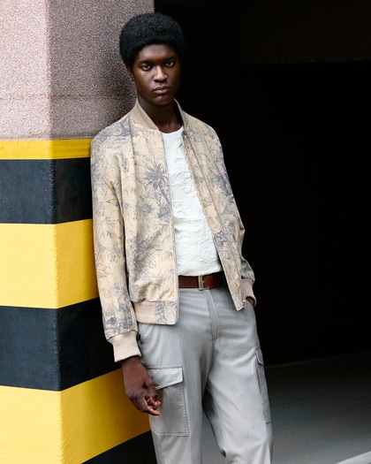 A man leans against a wall wearing a patterned beige bomber jacket, a textured white t-shirt, and light grey cargo pants.